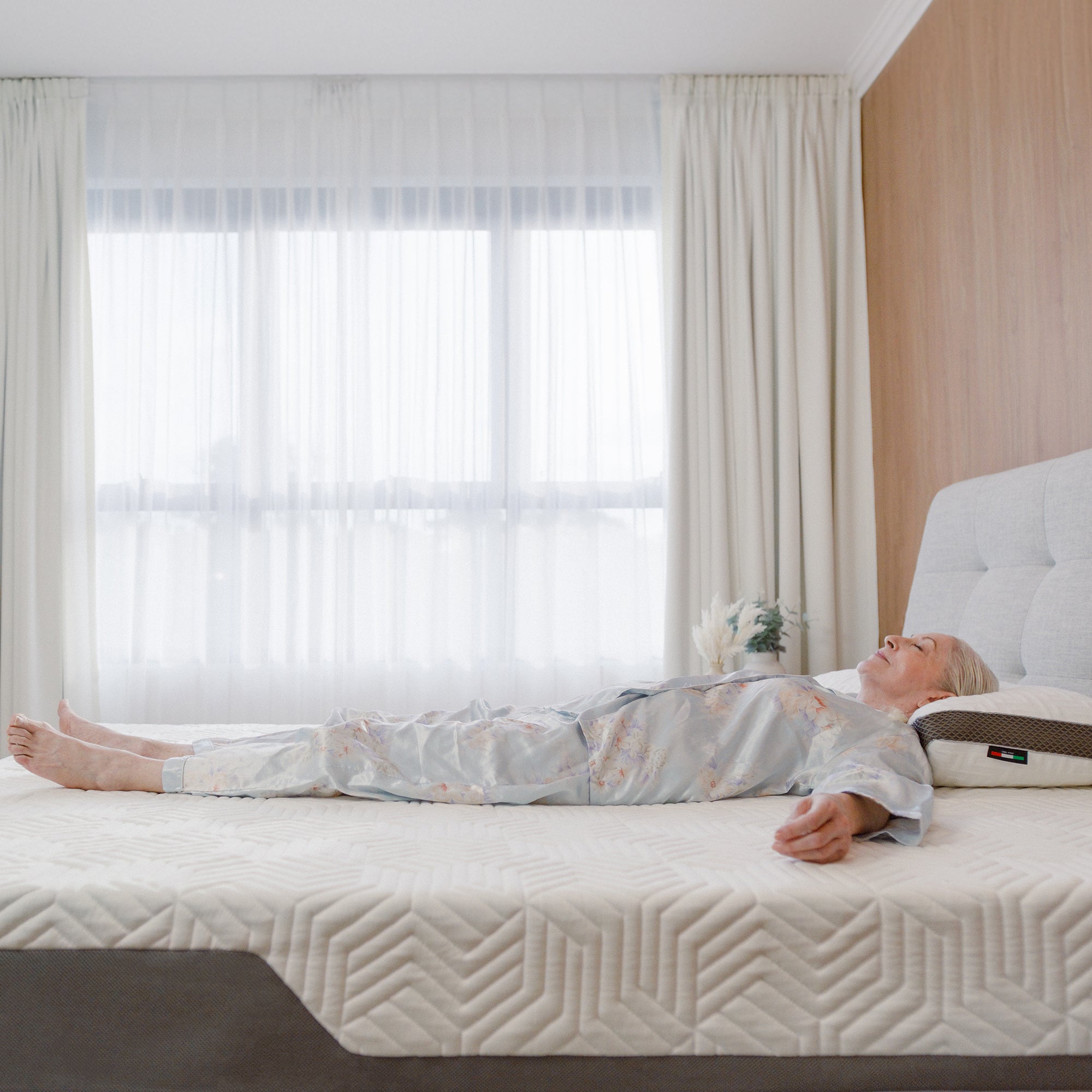 A person lying on a Valmori Hybrid Mattress Ⅱ (Medium) bed in a well-lit room with large windows and white curtains.