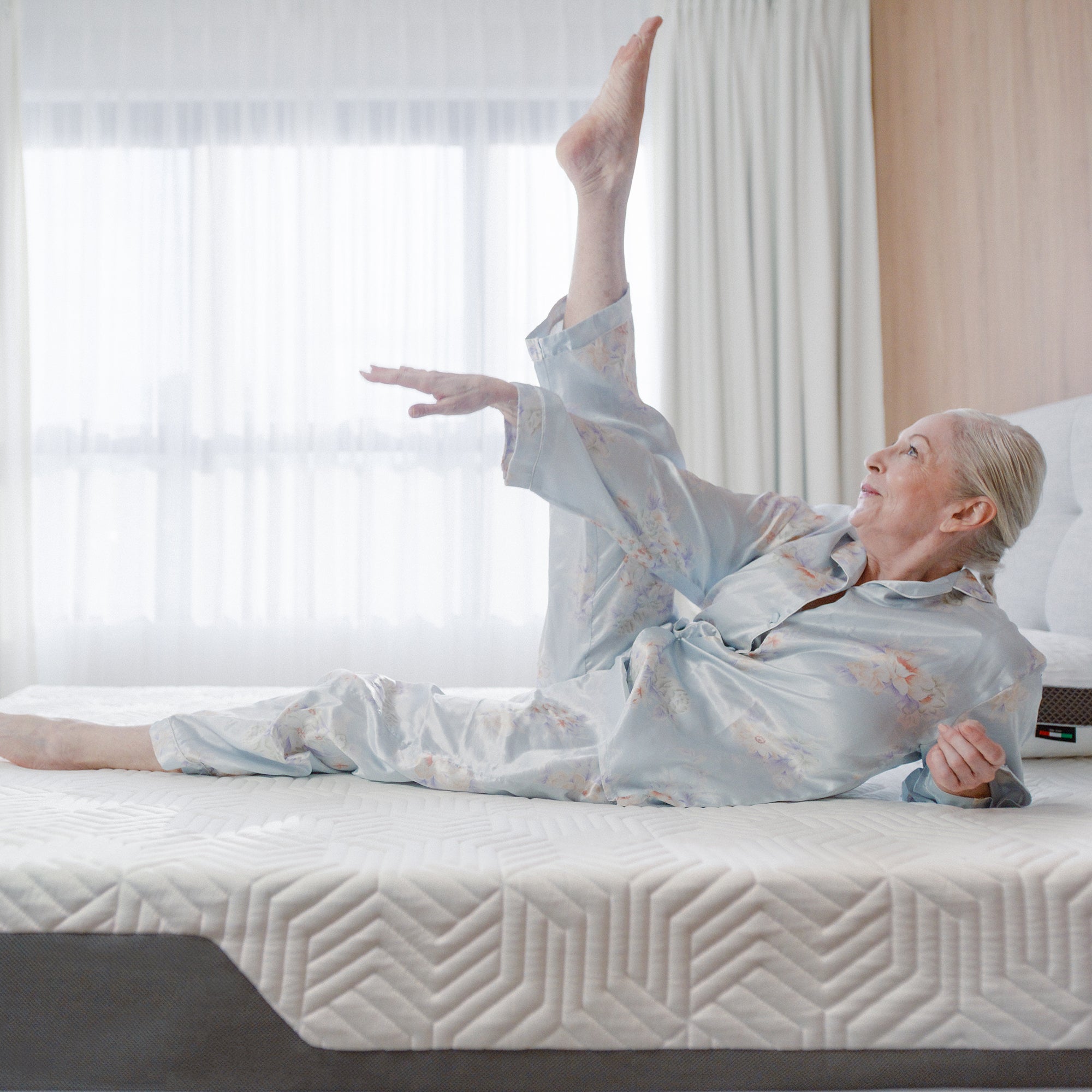 A woman wearing light-colored pajamas lying on a Valmori bed with one leg raised high in the air.